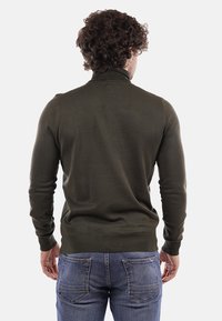 Maglione verde scuro con colletto alto, maniche lunghe e polsini e orlo a costine. Indossato con jeans blu; texture liscia e design aderente.