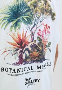 Hvid t-shirt med et livligt botanisk tryk af forskellige planter, tekst "BOTANICAL MIRACLE," og "GALLERY" logo i sort.