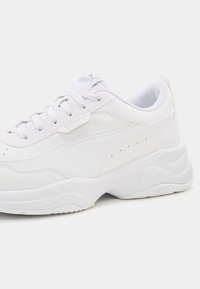 Baskets athlétiques blanches avec une tige en cuir lisse, embout arrondi, design à lacets et semelle intermédiaire amortie. Logo subtil et perforations présentes.