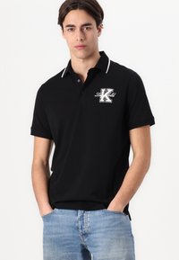 Černé polo tričko s bílým lemem, vybavené logem na hrudi. Vyrobeno z hladkého materiálu, krátké rukávy a klasický design límce.