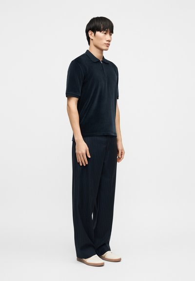 Μπλε βελούδινο πόλο πουκάμισο με κοντά μανίκια, συνδυασμένο με σκούρο ριγέ παντελόνι με ευρύ πόδι και ανοιχτά κρεμ παπούτσια.