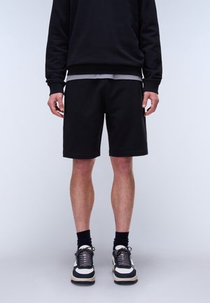 Man draagt een zwart shirt met lange mouwen, zwarte korte broek, zwarte sokken en zwart-witte sneakers, staand tegen een effen witte achtergrond.