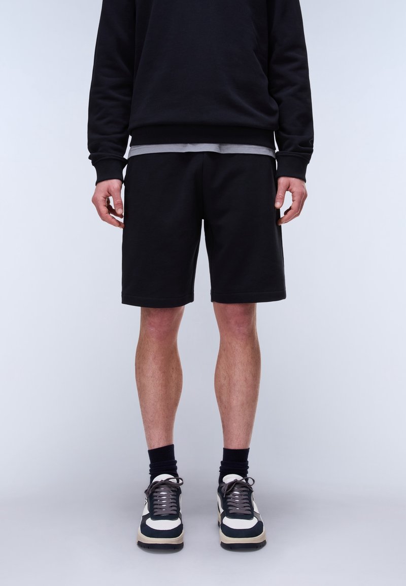 Homme portant une chemise à manches longues noire, un short noir, des chaussettes noires et des baskets noir et blanc, debout devant un fond blanc uni.