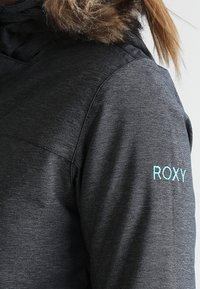 Manche de veste gris foncé avec le logo "ROXY" bleu clair brodé, capuche doublée de fourrure, et cheveux blonds attachés en arrière.