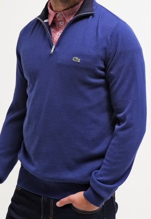 Homme portant un pull bleu à fermeture éclair quart de zip par-dessus une chemise rose boutonnée, une main dans la poche d'un jean foncé.