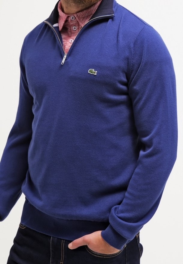 Homme portant un pull bleu à fermeture éclair quart de zip par-dessus une chemise rose boutonnée, une main dans la poche d'un jean foncé.