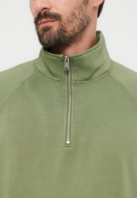 Άνδρας με γενειάδα που φοράει ελιάς πράσινο πουλόβερ με φερμουάρ στο ένα τέταρτο, εμφανίζοντας το ντεκολτέ και το άνω μέρος του στήθους έναντι απλού φόντου.