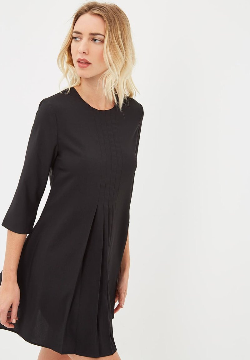 Mujer rubia vistiendo un vestido negro de mangas 3/4, con un frente plisado, mirando hacia su izquierda contra un fondo blanco liso.