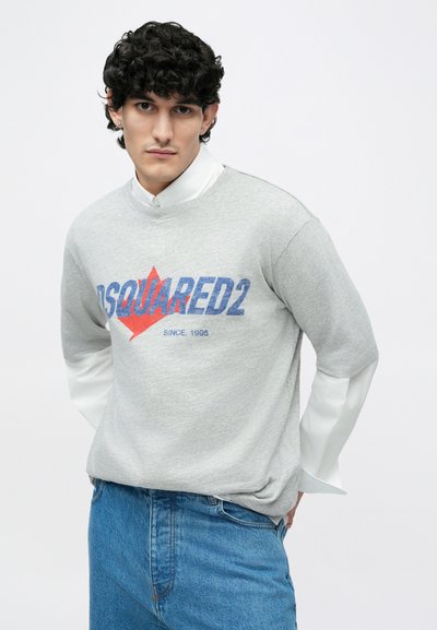 Grauer Pullover mit blauem und rotem "DSQUARED2"-Logo, kombiniert mit einem weißen Hemd darunter. Trägt dazu eine blaue Jeans.