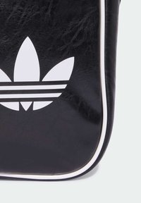 Melnas sintētiskās ādas soma ar baltām maliņām, kurai ir liels balts Trefoil logotips un trīs horizontālas melnas svītras apakšā.