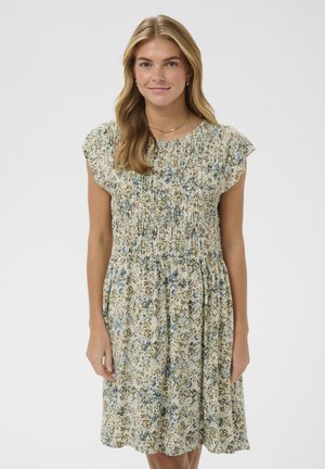 Žena s dlhými blond vlasmi, oblečená v krátkomá rukáve, kvetinových šatách po kolená s nariaseným živôtikom na jednoliatom svetlom pozadí.