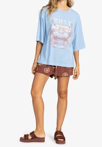 Mujer vistiendo una camiseta de gran tamaño de color azul claro con un gráfico de sol y palmera, pantalones cortos marrones con estampado y sandalias de dedo marrones.