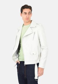 Giubbotto da motociclista in pelle bianca con tasche con cerniera, colletto a risvolto e vita con cintura. Indossato sopra un maglione a righe verde chiaro.