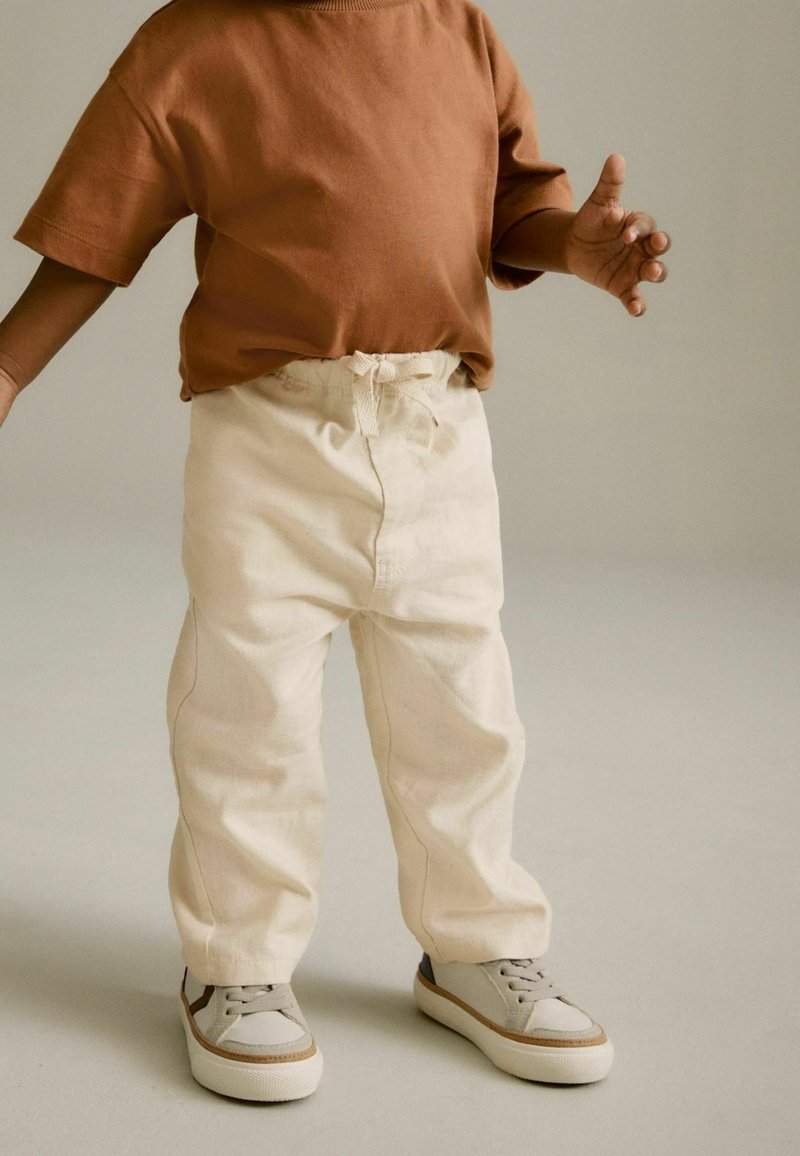 Tout-petit portant un t-shirt à manches longues marron, un pantalon couleur crème avec cordon de serrage, et des baskets beiges, debout sur un sol gris clair.