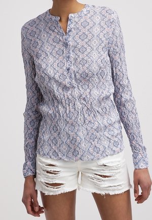 Nainen, joka pukeutunut pitkähihainen kuvioitu paita, jossa on napit, ja valkoiset kulutetut farkkushortsit.