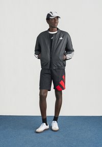 Zwarte bomberjack met witte strepen, zwarte korte broek met rode strepen en witte sneakers met beige zolen. Het model draagt ook een pet.