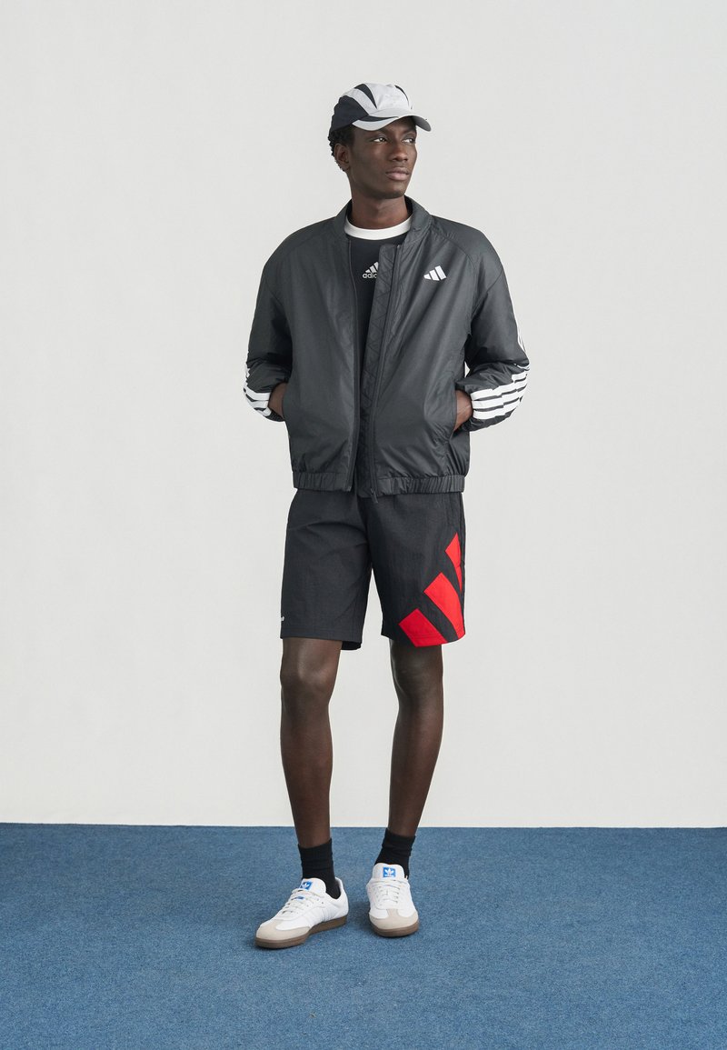 Schwarze Bomberjacke mit weißen Streifen, schwarze Shorts mit roten Streifen und weiße Turnschuhe mit hellbraunen Sohlen. Das Model trägt außerdem eine Mütze.