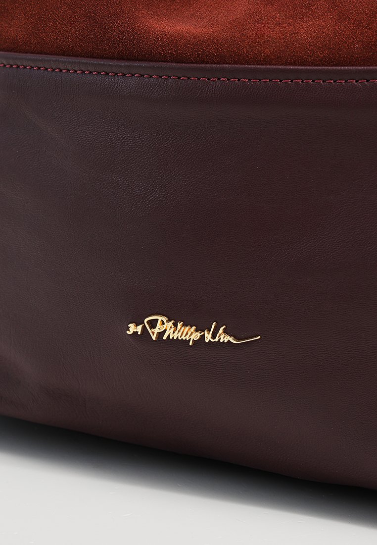 Burgundowa skórzana torba charakteryzuje się gładką teksturą i złotym logo podpisowym. Projekt zawiera kontrastujący zamszowy materiał na górze.