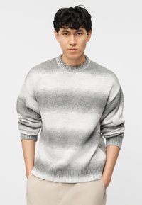 Νεαρός άνδρας με κοντά σγουρά μαλλιά, που φοράει γκρι και λευκό ριγέ πουλόβερ και μπεζ παντελόνια, με τα χέρια στις τσέπες, ουδέτερη έκφραση, λευκό φόντο.