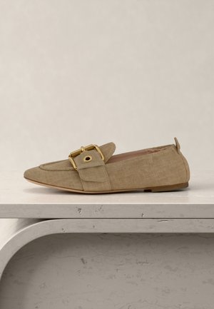 Beige fabric loafer with pointed toe, side flap, large gold buckle, and flat sole on textured light gray surface.