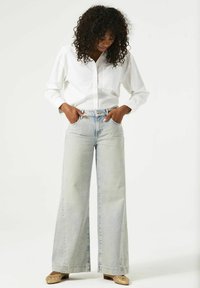 Jeans de mezclilla de tiro alto y pierna ancha en azul claro, combinados con una camisa blanca abotonada. Los jeans presentan un lavado descolorido y costuras visibles.
