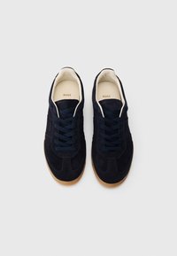 Σκούρα μπλε σουέτ αθλητικά παπούτσια με σόλα από καουτσούκ χρώματος τσίχλας, με στρογγυλά κορδόνια και διακριτική ραφή σε τόνους στο πάνω μέρος. Φέρουν την επιγραφή "BOSS" εσωτερικά.