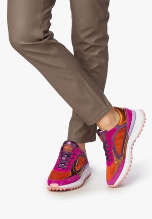 Zapatilla deportiva morada y rosa con detalles naranjas, de ante y material sintético, lengüeta acolchada y suela texturizada. Combinada con pantalones ajustados color beige.