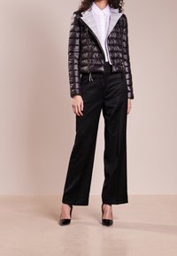 Femme portant une doudoune noire brillante par-dessus une chemise blanche à col, un pantalon large noir et des escarpins noirs à bout pointu, se tenant sur un sol beige.