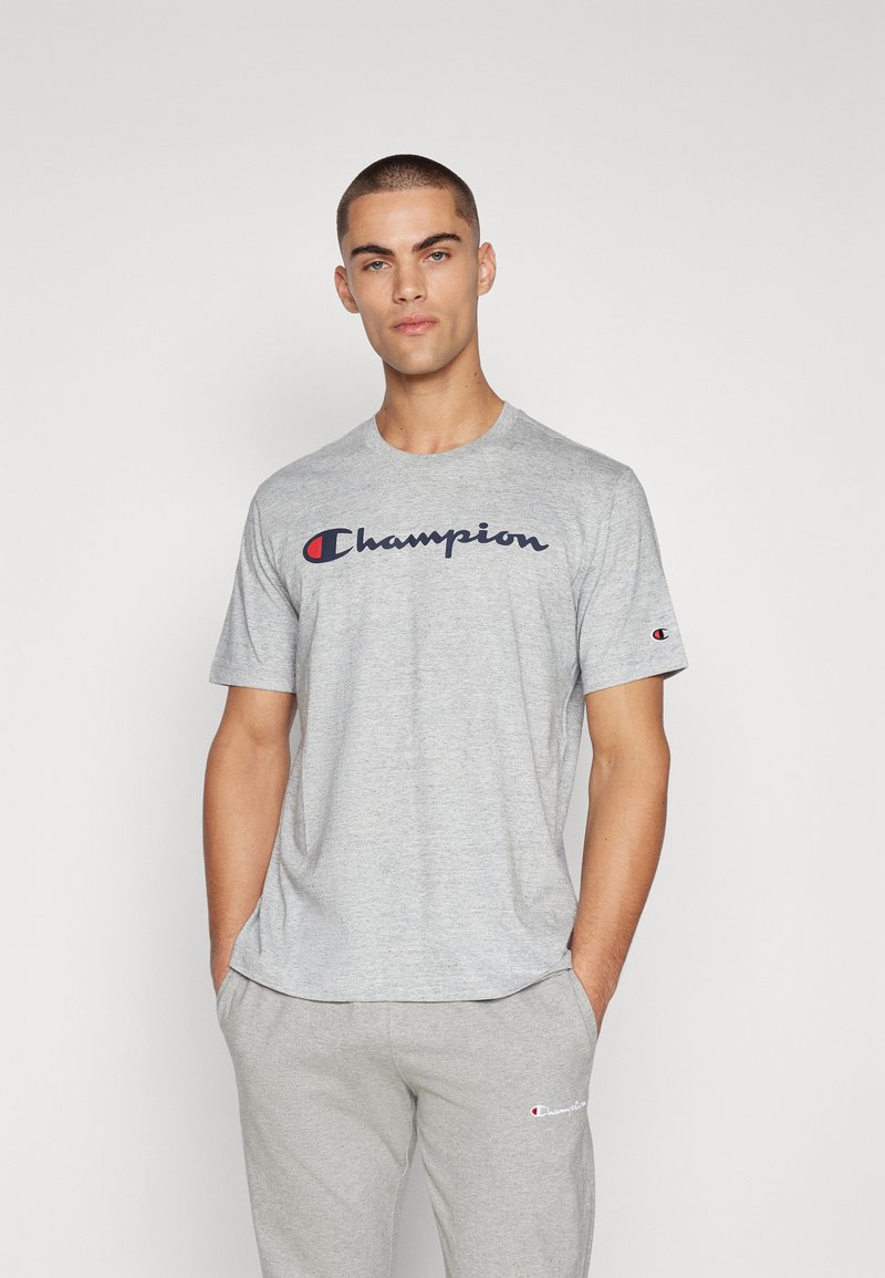 Graues Baumwoll-T-Shirt mit kurzen Ärmeln, das ein navyblaues und rotes "Champion"-Logo über der Brust trägt. Lässige Passform, weicher Stoff.