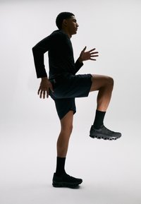Sportliche Schuhe in Schwarz mit einer strukturierten Sohle, kombiniert mit schwarzen Shorts und einem Langarmtop. Person in einer Laufpose.