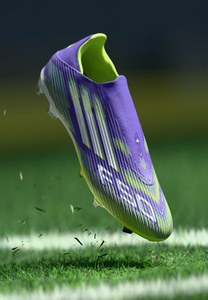 Purple and green soccer cleat with a textured surface, sharp stripes, and a low-cut design, elevated above grass with dirt particles.