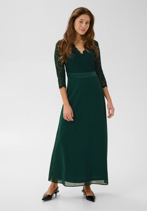 Une femme porte une robe longue jusqu’au sol, vert foncé, avec des manches en dentelle et un décolleté en V, assortie à des chaussures noires à talons, sur un fond de studio uni.