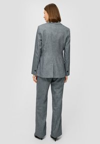 Grijs maatwerkpak met een gestructureerde afwerking, voorzien van een enkele split aan de achterkant en lange broeken. Er zijn decoratieve knopen op de mouwen.