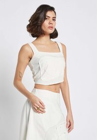 Weißes Cropped-Top mit quadratischem Ausschnitt, breiten Trägern und strukturiertem Stoff, der eine auffällige Vordernaht aufweist. Kombiniert mit einem passenden Rock.