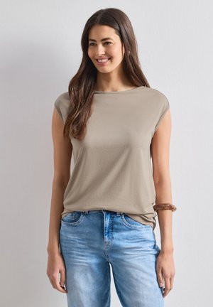 Frau mit langen braunen Haaren, die ein beiges kurzärmeliges Oberteil und hellblaue Jeans trägt, steht und lächelt vor einer schlichten hellen Wand.