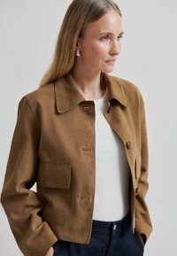 Barna velúr dzseki, galléros nyakkal, két elülső zsebbel és egy gombbal záródik. Fehér ing felett viselve.