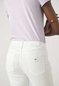 Bílé džíny z denimu se dvěma zadními kapsami. Na zadním pásku se nachází látková nášivka s nápisem "ARMANI EXCHANGE." Vrchní část je světle fialová z bavlny.