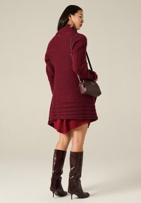 Cappotto a maglia bordeaux con colletto alto, motivo testurizzato e accenti a coste; abbinato a un mini abito bordeaux e stivali lucidi fino al ginocchio.
