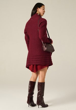Cappotto a maglia bordeaux con colletto alto, motivo testurizzato e accenti a coste; abbinato a un mini abito bordeaux e stivali lucidi fino al ginocchio.