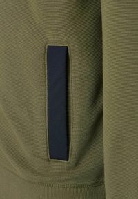 Olivgrüner Sweatstoff mit gerippter Textur, ausgestattet mit einer navyblauen, gesteppten Taschenapplikation auf der linken Seite.