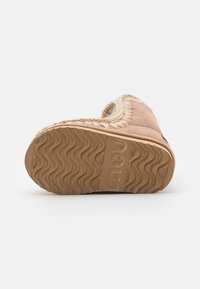 Botte en brun clair, duveteuse, avec une semelle en caoutchouc texturée présentant un motif en vagues. Couture décorative le long du bord supérieur. Logo circulaire intégré.