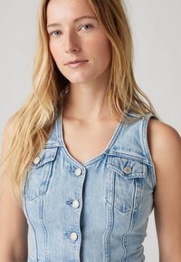 Svetle modré džíny bez rukávů s výstřihem do V, předními knoflíky a kapsami na prsou. Má přiléhavý střih a mírnou texturu.