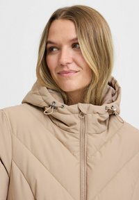 Giacca a vento beige con cappuccio, caratterizzata da un motivo trapuntato diagonale, chiusura frontale con cerniera e cordini regolabili sul cappuccio.