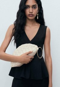 Bílá tkaná kabelka typu clutch se zlatým kroužkem, držená před modelem v černém topu s peplum designem a výstřihem do V.