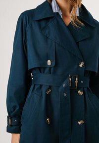 Manteau trench bleu marine, à double boutonnage, col large, poignets ajustables et taille ceinturée. Tissu lisse avec un subtil éclat.