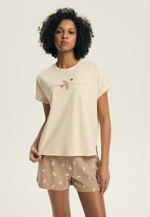 Kobieta o kręconych włosach, ubrana w beżowy t-shirt z grafiką ptaka oraz pasujące szorty z małymi wzorami ptaków, stojąca na jednolitym tle.