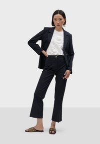 Blazer azul marino sobre camiseta blanca con números en relieve, acompañado de pantalones de cintura alta azul marino y sandalias negras y doradas con estampado.