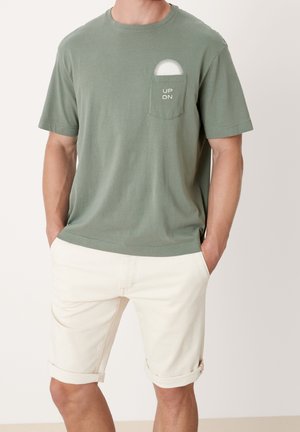 Grünes Baumwoll-T-Shirt mit kurzen Ärmeln, ausgestattet mit einer Tasche mit einer weißen Sonne und dem Text "UP DN." Kombiniert mit hochgekrempelten hellbeigen Shorts.