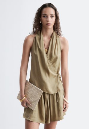 Jeune femme aux cheveux ondulés portant un top cache-cœur sans manches vert olive et une jupe assortie, tenant une pochette beige texturée.