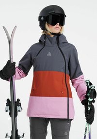 Ski bunda v šedej, červenej a ružovej farbe s kapucňou, zipsovými vreckami a detailom loga. Čierna prilba a lyžiarske okuliare, drží lyže a paličky.